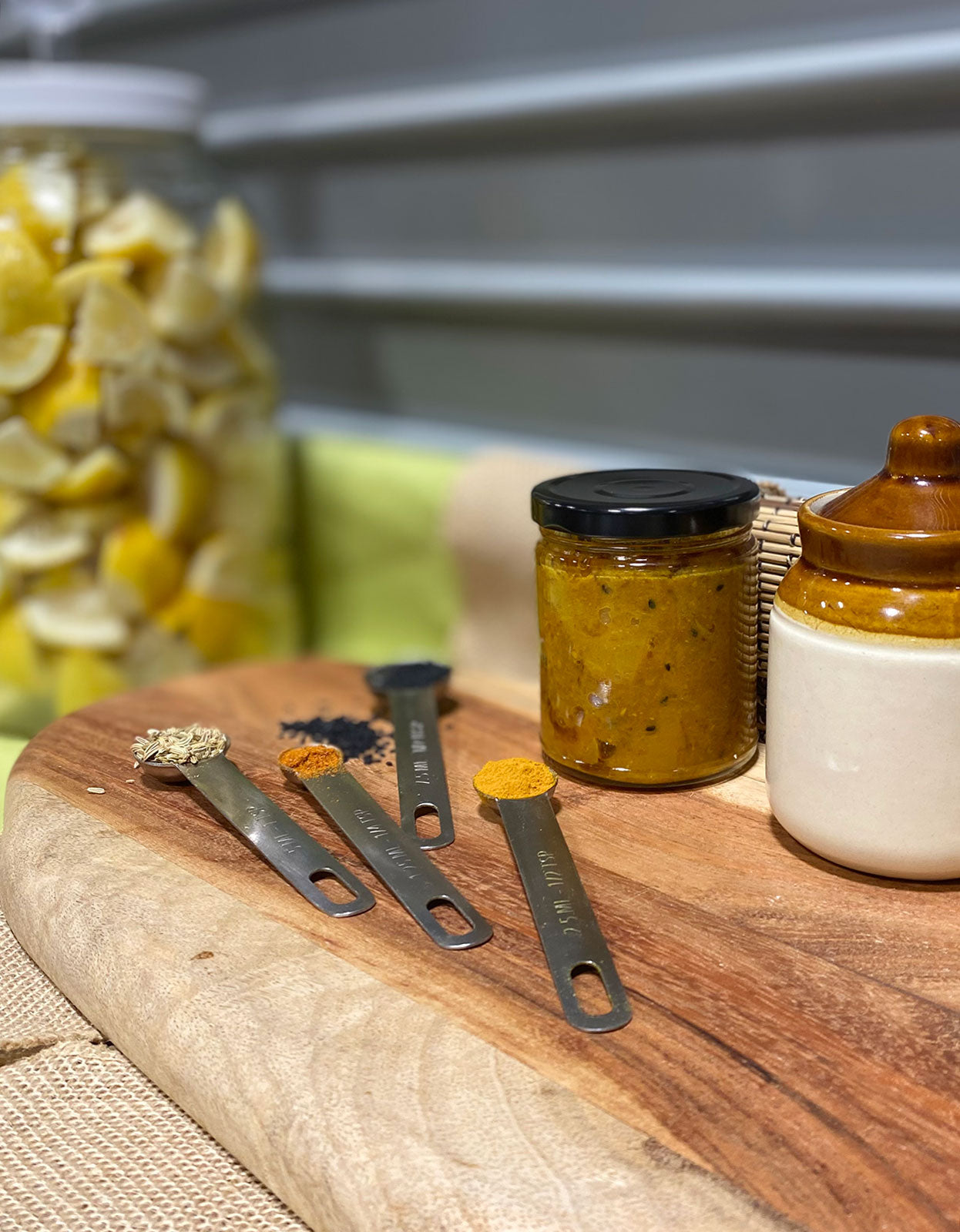 a wooden board with steel measuring spoons full of spices next to a jar of Nimbu ka Achar, an indian lemon pickle