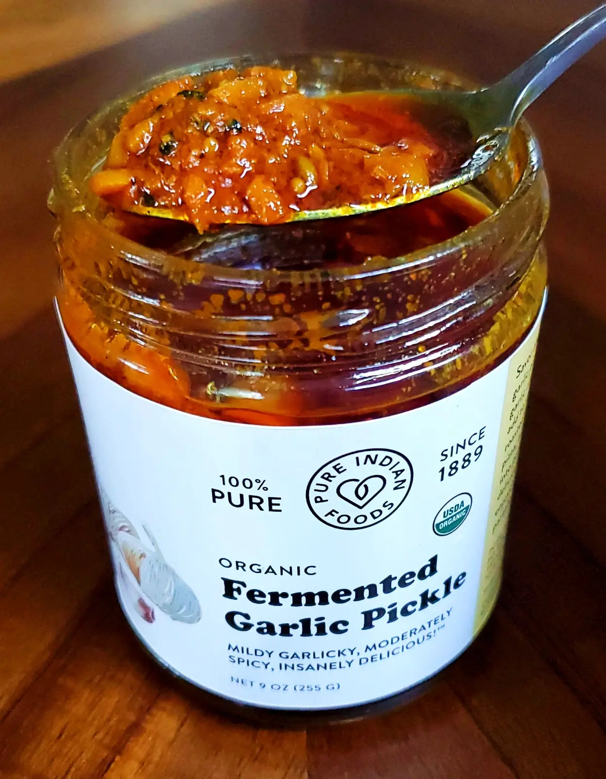 An open jar of Organic Fermented Garlic Pickle from Pure Indian Foods, with a spoonful of spicy pickled garlic achaar held above the open glass jar. The Indian Pickle is red, oily, and full of chopped garlic and fiery, aromatic spices.