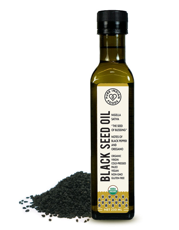 Bottle of Pure Indian Foods Cold Pressed Black Seed Oil displayed next to a mound of organic black cumin seeds. Label says it's extracted from Nigella Sativa, "the seed of blessing," and that it's organic, Virgin, cold-pressed, Paleo, vegan, non-gmo, organic, and gluten-free.