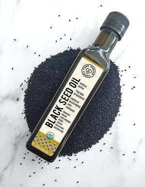 Bottle of Pure Indian Foods Cold-Pressed Black Seed Oil artfully displayed on a bed of black cumin seeds.