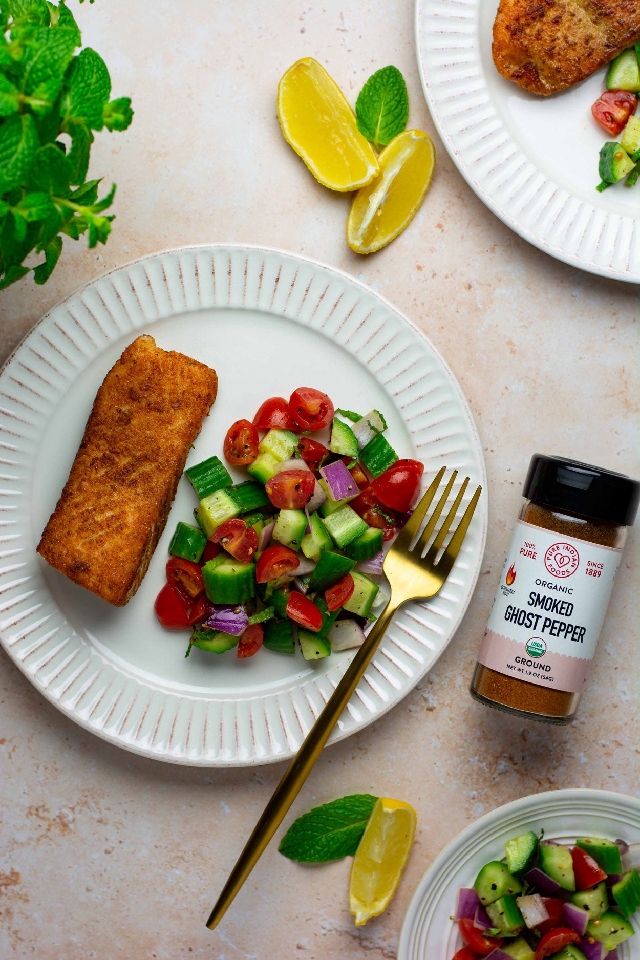 A birds eye view of a white dinner plate topped with a seared salmon steak and a salad of fresh veggies next to a jar of the organic smoked ghost pepper powder from Pure Indian Foods