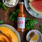 1 bottle of our Pure Indian Foods KICK Hot Sauce, a certified organic Mango Ghost pepper hot sauce. Displayed on a table with sliced cantaloupe and watermelon, cheese and honey.