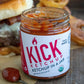 1 open jar of Pure Indian Foods KICK Ketchup, an organic ketchup. A heaping spoonful of ketchup is to the left of the jar, and a yummy looking burger with homemade onion rings is behind it.