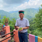 Sandeep standing on a balcony in India with a stunning forested mountain view behind him, holding freshly picked Myrobalan fruit in his hands.