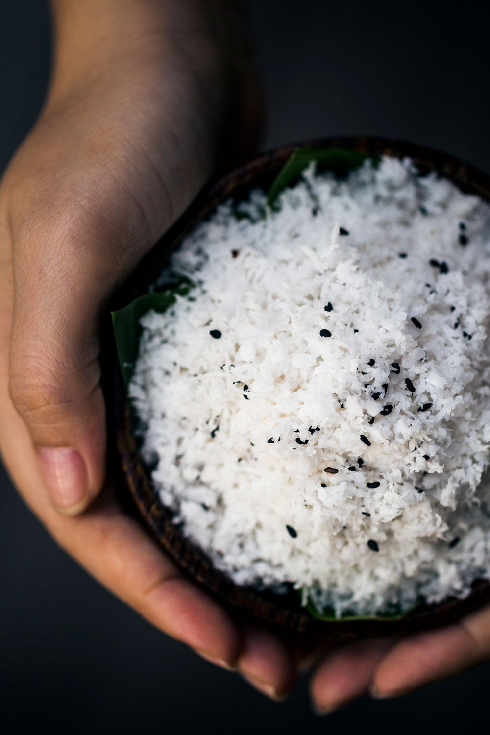 Close up of hands wrapped around a bowl of shredded coconut and black sesame seeds