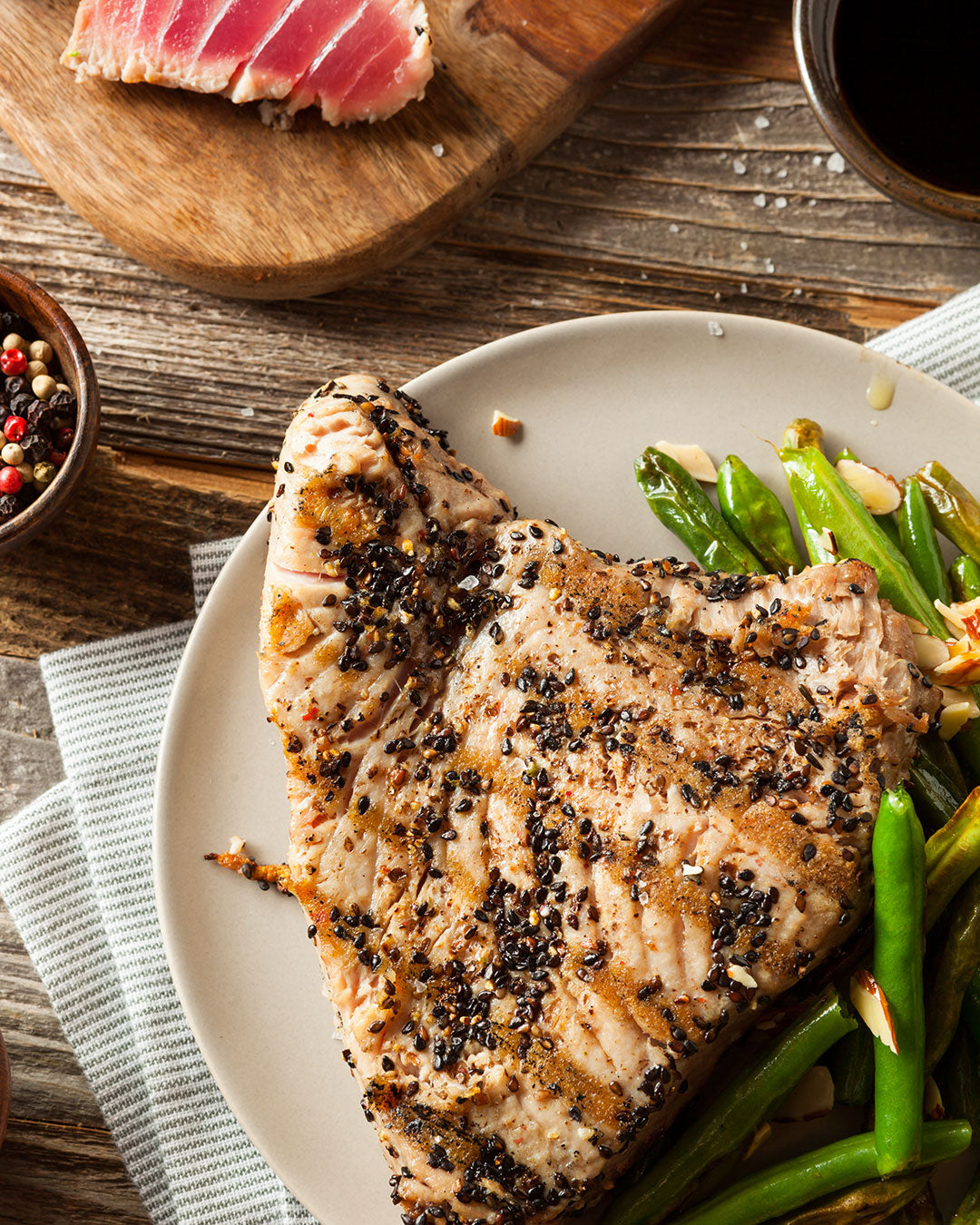 A grilled tuna steak coated in black sesame seeds with a soy glaze, served on a white plate with green beans and almond slivers.