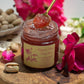 Open jar of Organic Rose Petal Jam, artfully displayed on a live edge wooden charcuterie board next to roses.  Someone holds a small spoonful of the rose jam