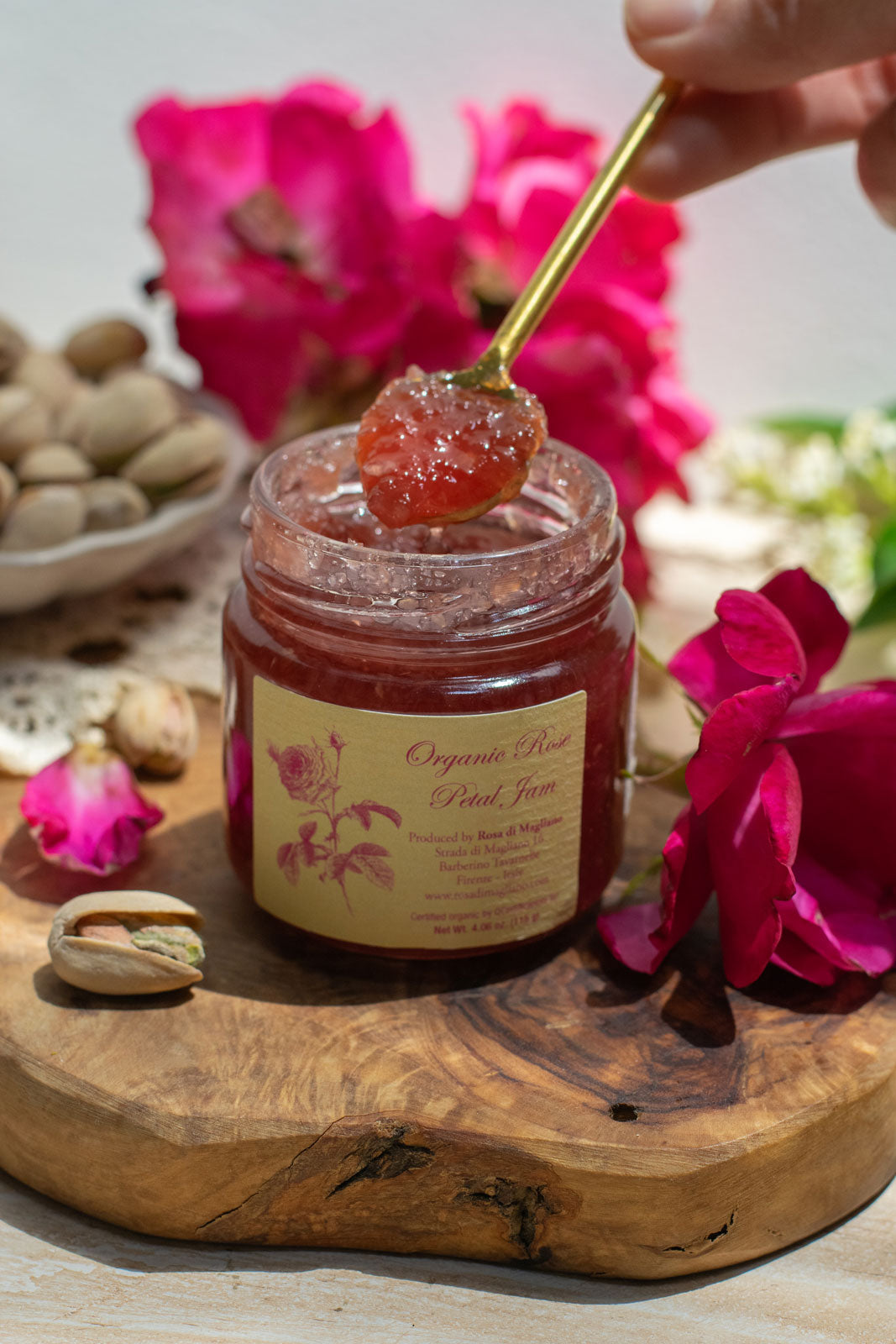Open jar of Organic Rose Petal Jam, artfully displayed on a live edge wooden charcuterie board next to roses.  Someone holds a small spoonful of the rose jam
