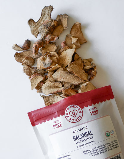 An open package of Organic Galangal Slices (dried) from Pure Indian Foods spilling out onto a white surface.