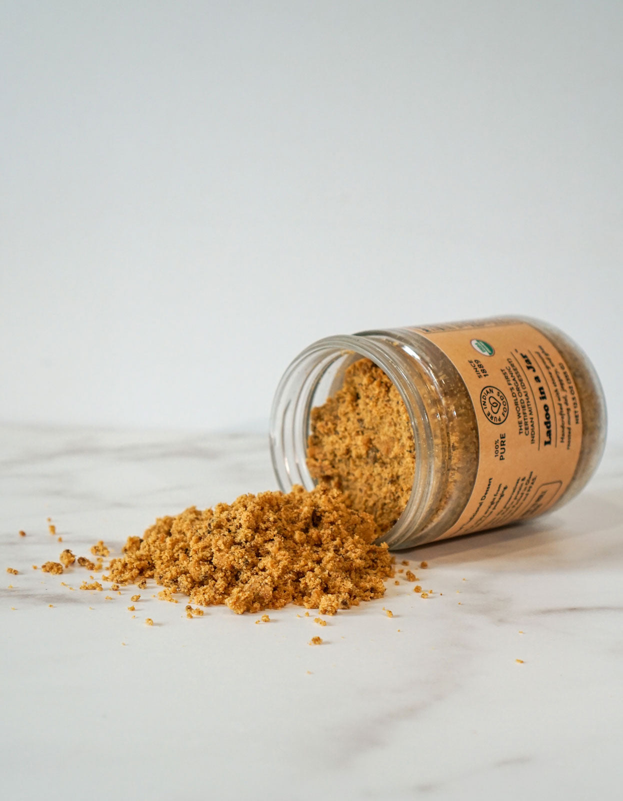 An open jar of Pure Indian Foods Organic Ladoo in a Jar spilling onto a marble counter.