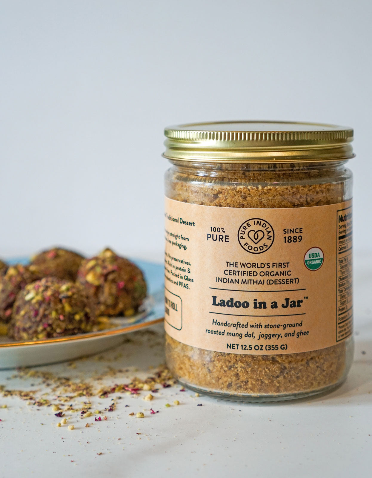 A certified organic ladoo mix that already includes ghee and jaggery, featured in a glass jar in front of a plate of beautiful Ladoos.