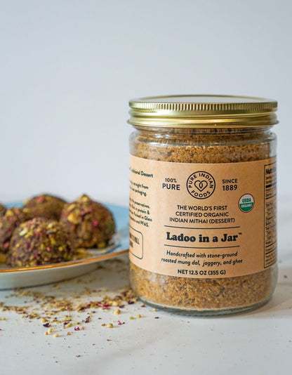 A certified organic ladoo mix that already includes ghee and jaggery, featured in a glass jar in front of a plate of beautiful Ladoos.