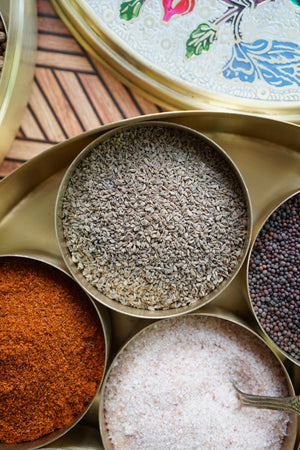 A brass masala dabba filled with various spices, focused on a container of whole carom seeds (also known as ajwain seed).