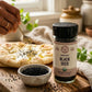 A jar of organic kalonji on a wooden board next to a bowl of black seeds artfully arranged in front of someone's hand sprinkling the seeds on a flatbread.