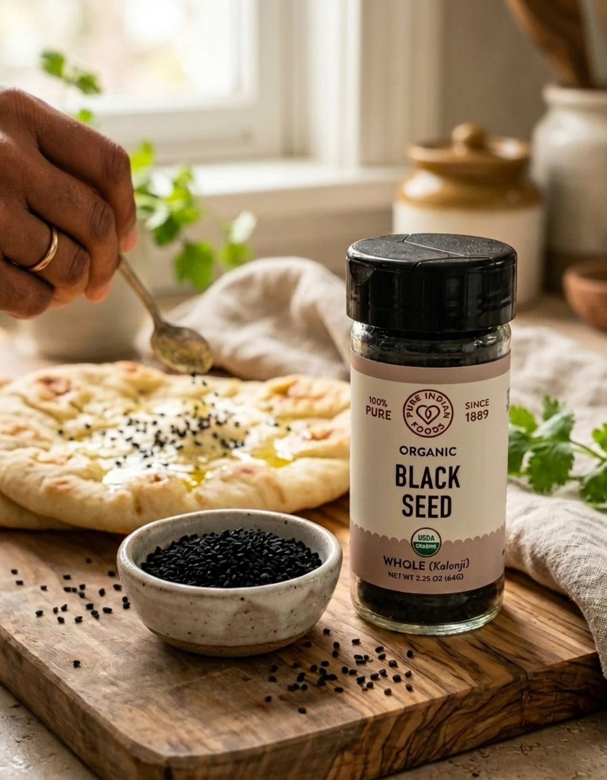 A jar of organic kalonji on a wooden board next to a bowl of black seeds artfully arranged in front of someone's hand sprinkling the seeds on a flatbread.
