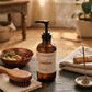 An amber glass pump bottle labeled 'GREEK BEAUTY CALMING SCALP & BODY OIL' sits on a stone surface in a warm, rustic bathroom or spa setting with a wooden brush, incense holder, and steam in the background.