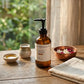 A bottle of Gheek Beauty Cooling Massage oil with a black pump, set on a rustic wooden table with a beige cloth, small bowls, and a copper dish with flowers, with a soft-focus background of green foliage and a curtain.