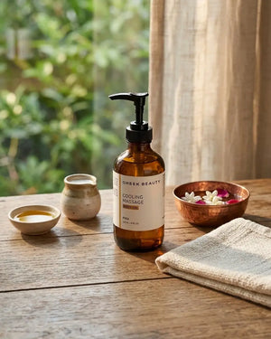 A bottle of Gheek Beauty Cooling Massage oil with a black pump, set on a rustic wooden table with a beige cloth, small bowls, and a copper dish with flowers, with a soft-focus background of green foliage and a curtain.