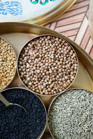 Whole coriander seed from Pure Indian Foods in a brass masala dabba surrounded by other organic spices.