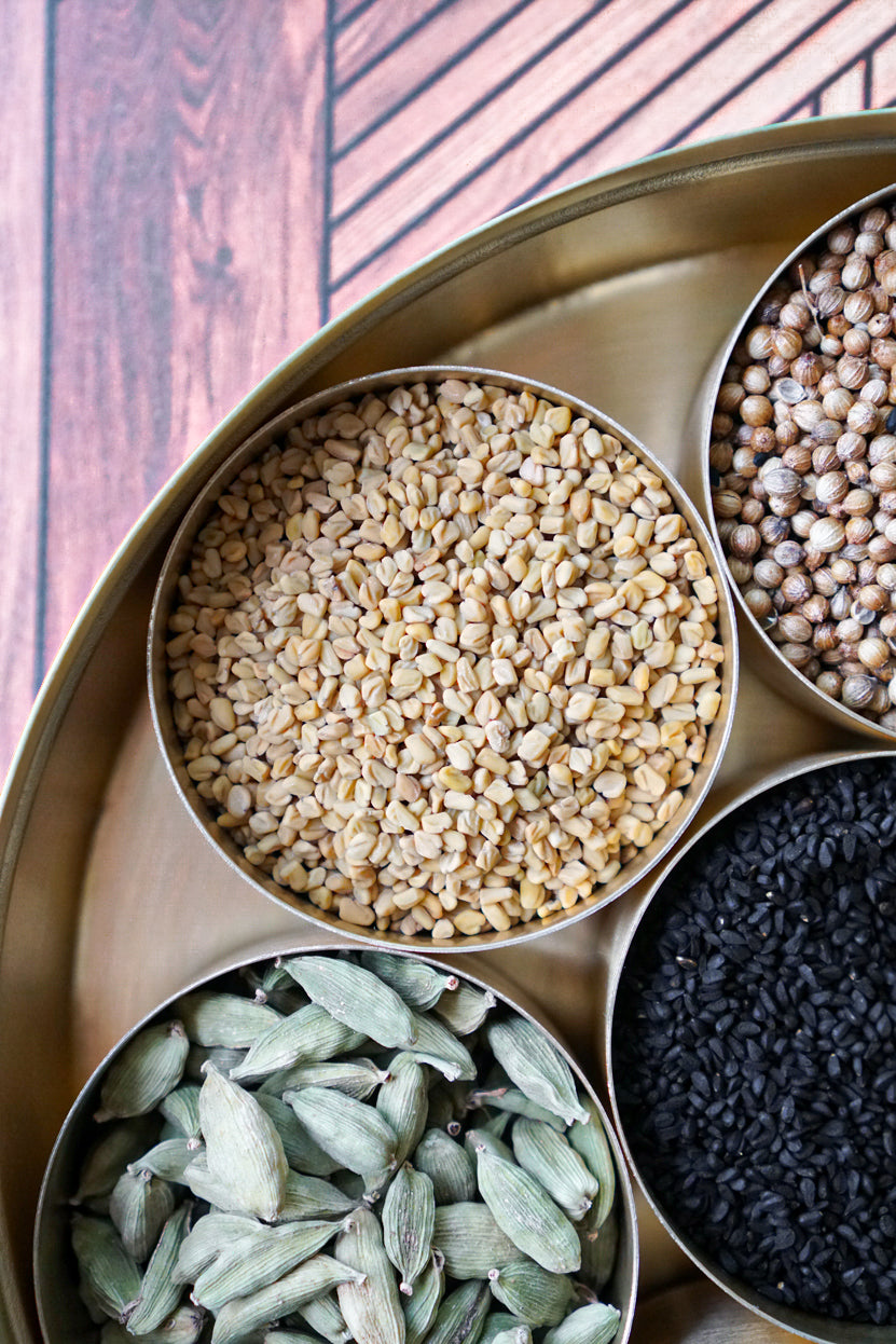 Whole, organic fenugreek seeds from Pure Indian Foods displayed in a brass masala dabba among other organic spices.