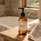 An amber glass pump bottle of Gheek Beauty Fortifying Face Wash sits on a rustic wooden bathroom counter next to a folded towel, with a blurred stone-tiled bathroom background.