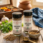Two jars of organic coriander seed spices on a rustic wooden table, surrounded by fresh cilantro, a small bowl of ground coriander with a spoon, a brass spoon, and a wooden spice box in the background.