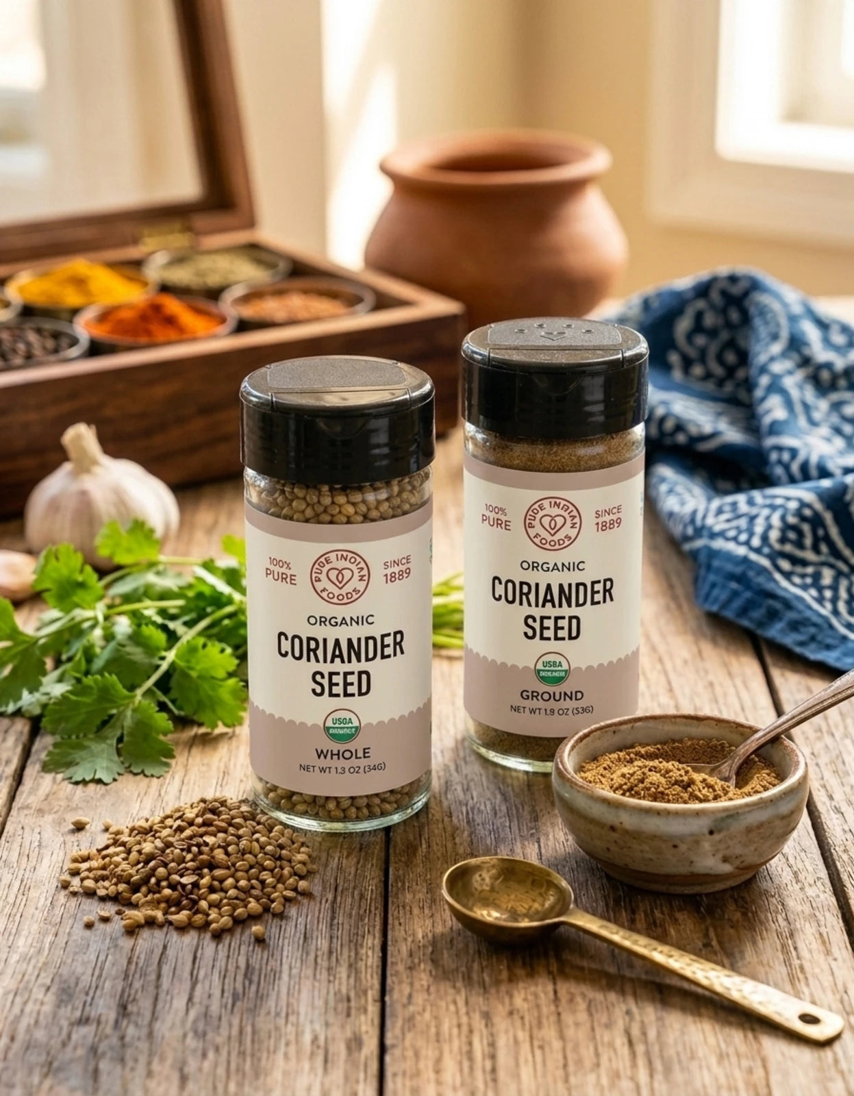 Two jars of organic coriander seed spices on a rustic wooden table, surrounded by fresh cilantro, a small bowl of ground coriander with a spoon, a brass spoon, and a wooden spice box in the background.