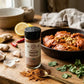 A bottle of organic tandoori masala seasoning blend on a wooden table next to a skillet full of tandoori chicken and surrounded by fresh herbs and spices