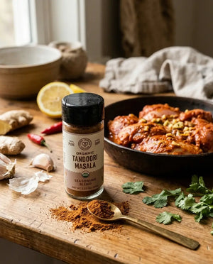 A bottle of organic tandoori masala seasoning blend on a wooden table next to a skillet full of tandoori chicken and surrounded by fresh herbs and spices