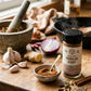 A bottle of Pure Indian Foods Organic Vindaloo Curry Seasoning Blend on a wooden counter surrounded by onions, ginger, garlic, and herbs