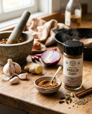 A bottle of Pure Indian Foods Organic Vindaloo Curry Seasoning Blend on a wooden counter surrounded by onions, ginger, garlic, and herbs