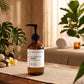 An amber glass pump bottle labeled GHEEK BEAUTY Rejuvinating Cleanser sits on a wooden table in a spa-like setting with plants, a towel, and warm ambient lighting.