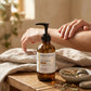 An amber glass pump bottle of Cheek Beauty Restorative Body Oil sits on a wooden table beside a person's arm receiving a massage, with a neutral beige background and spa props including a towel and a small dish of herbs.