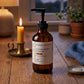 An amber pump bottle of Sandalwood Evening Body Oil from Gheek Beauty on a wooden table in the evening light next to a lit candle and warm lamp.