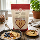 A stand-up pouch of Pure Indian Foods Sprouted Amaranth. It is displayed on a rustic wooden table next to a bowl of prepared amaranth porridge and a small dish of loose amaranth seeds. The background features a window, a potted basil plant, and a jar of almonds.