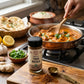 A bottle of Organic Tikka Masala seasoning on a wooden board in front a pan of chicken tikka masala cooking on a stove top.