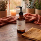 An amber glass pump bottle labeled 'GHEEK BEAUTY WARMING MASSAGE BODY OIL' stands on a wooden table in a warm, styled interior with a candle, plants, and draped fabric in the background.