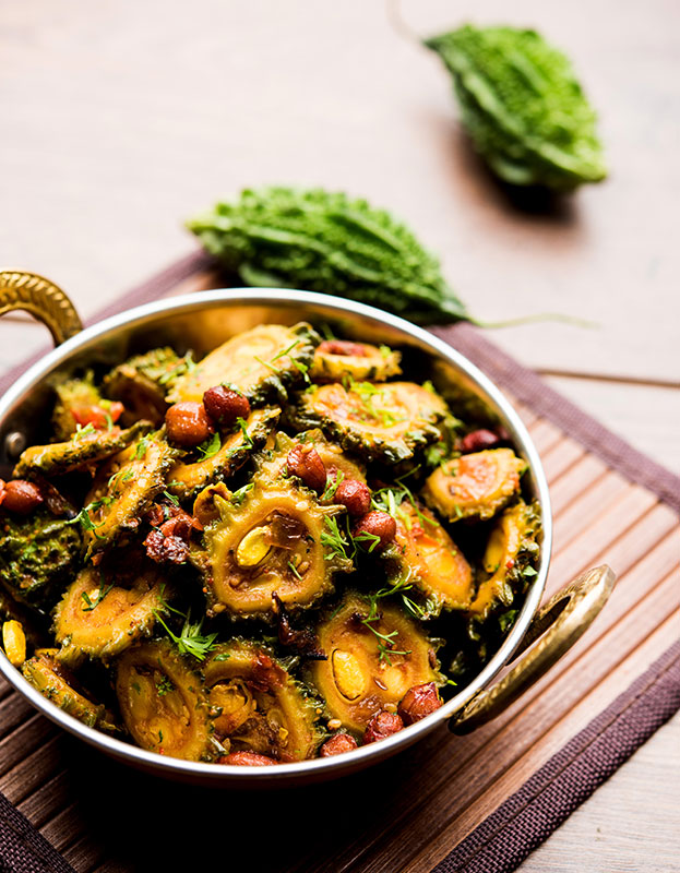 Indian dish made with rehydrated dried bitter melon slices, served in a brass and copper karahi serving bowl. Fresh, whole bitter melon is in the background.