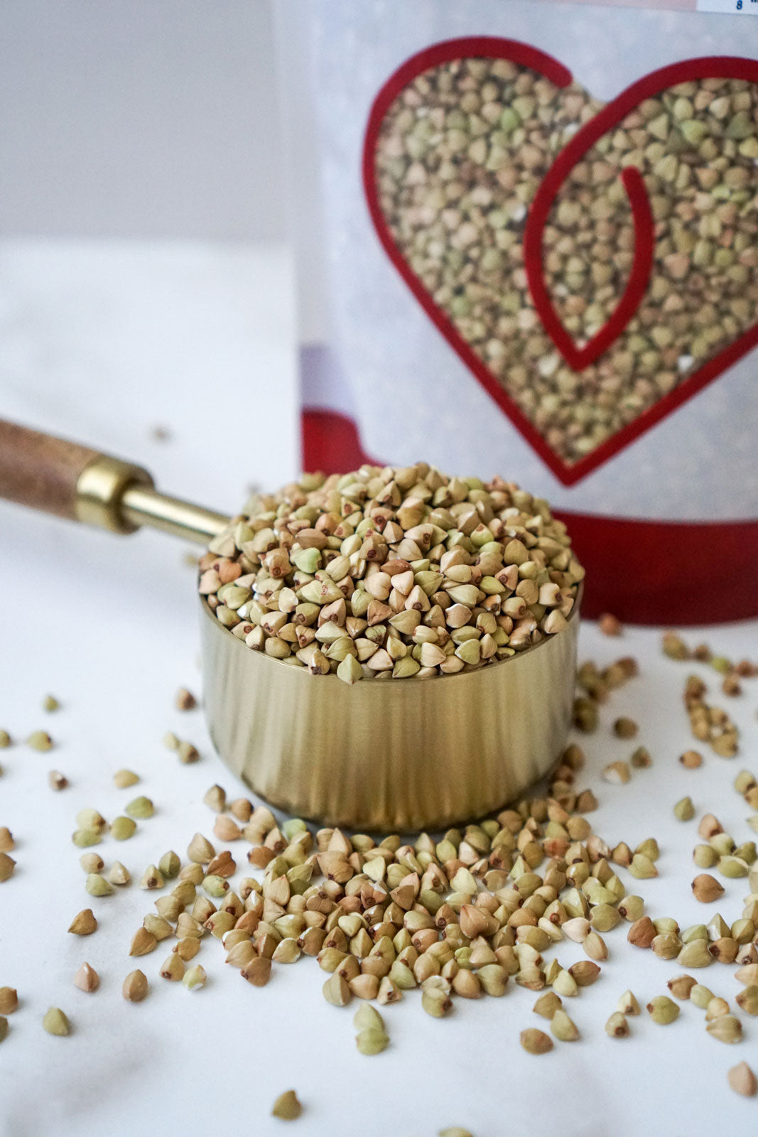 a brass measuring cup overflowing with organic whole buckwheat in front of the product bag from Pure Indian Foods