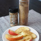 A jar of Pure Indian Foods Bombay Chaat Masala pictured next to a glass canister of the same spice mix that's been freshly ground. In front of both, a plate of sliced apples sprinkled with organic Chaat masala sits on a table.