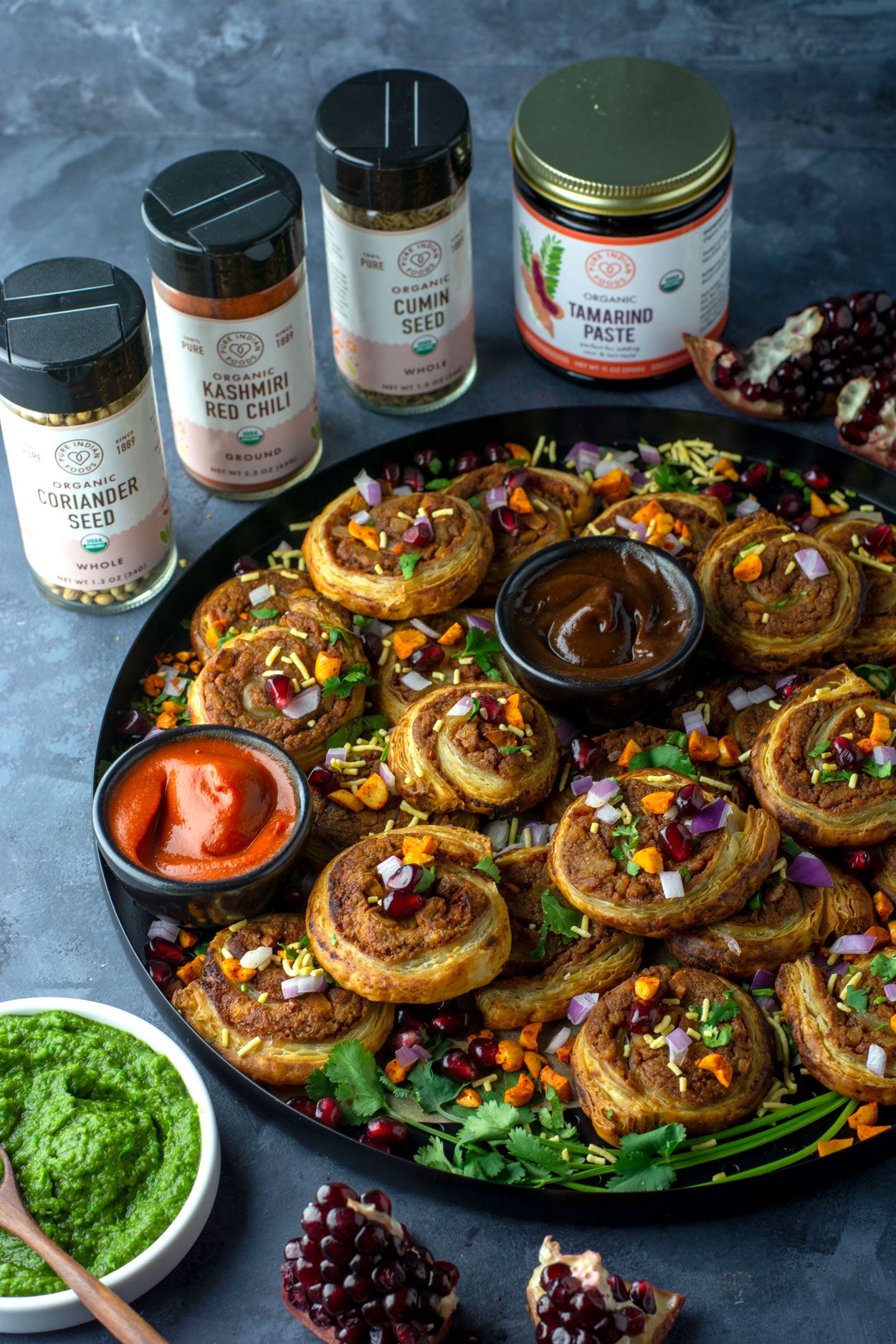 A tray of scrumptious pinwheels made with Pure Indian Foods products, including Organic Coriander Seed, Organic Kashmiri Chili Powder, Organic Cumin Seed, and Organic Tamarind Paste, all of which are displayed behind the pinwheels.