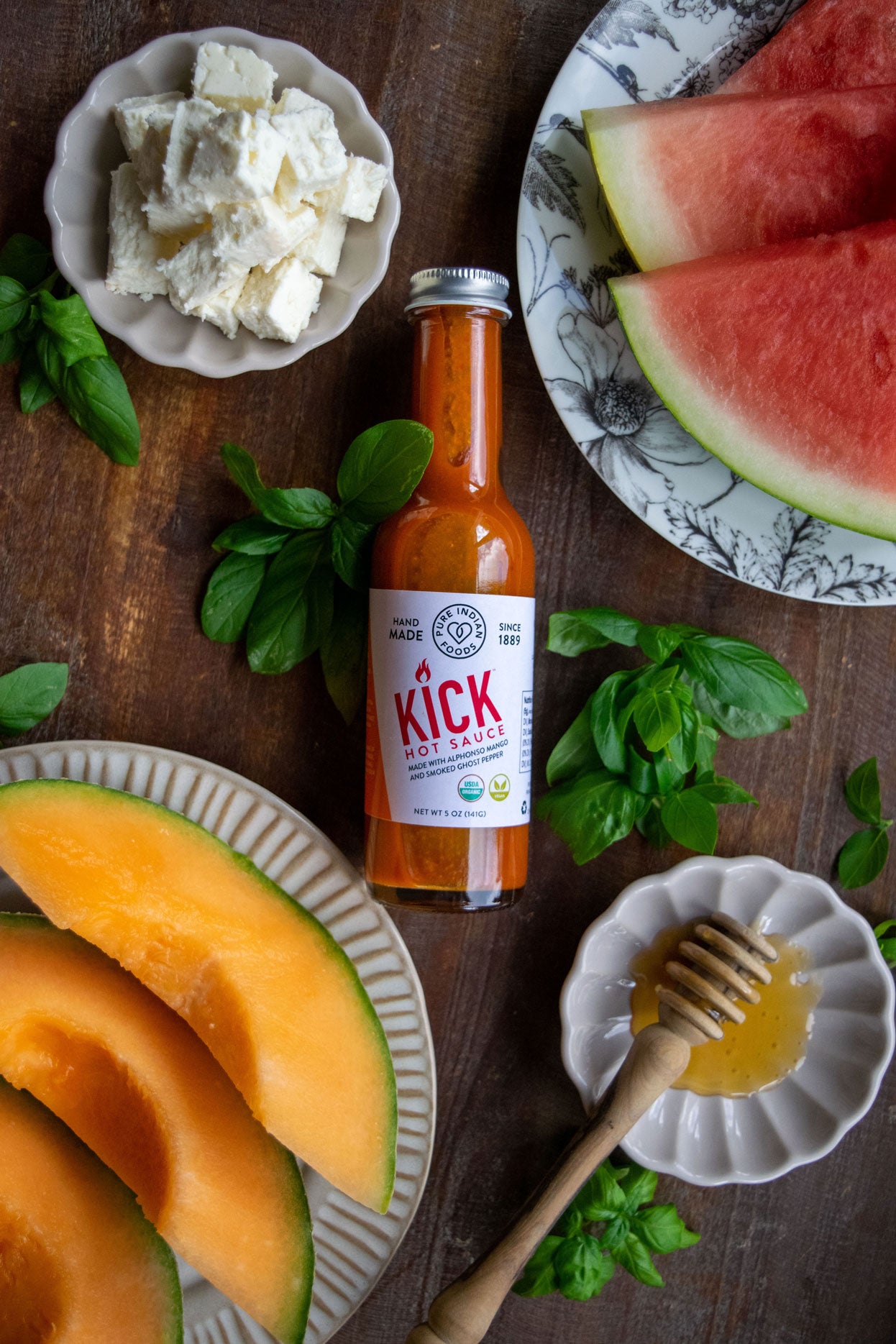 1 bottle of our Pure Indian Foods KICK Hot Sauce, a certified organic Mango Ghost pepper hot sauce. Displayed on a table with sliced cantaloupe and watermelon, cheese and honey.