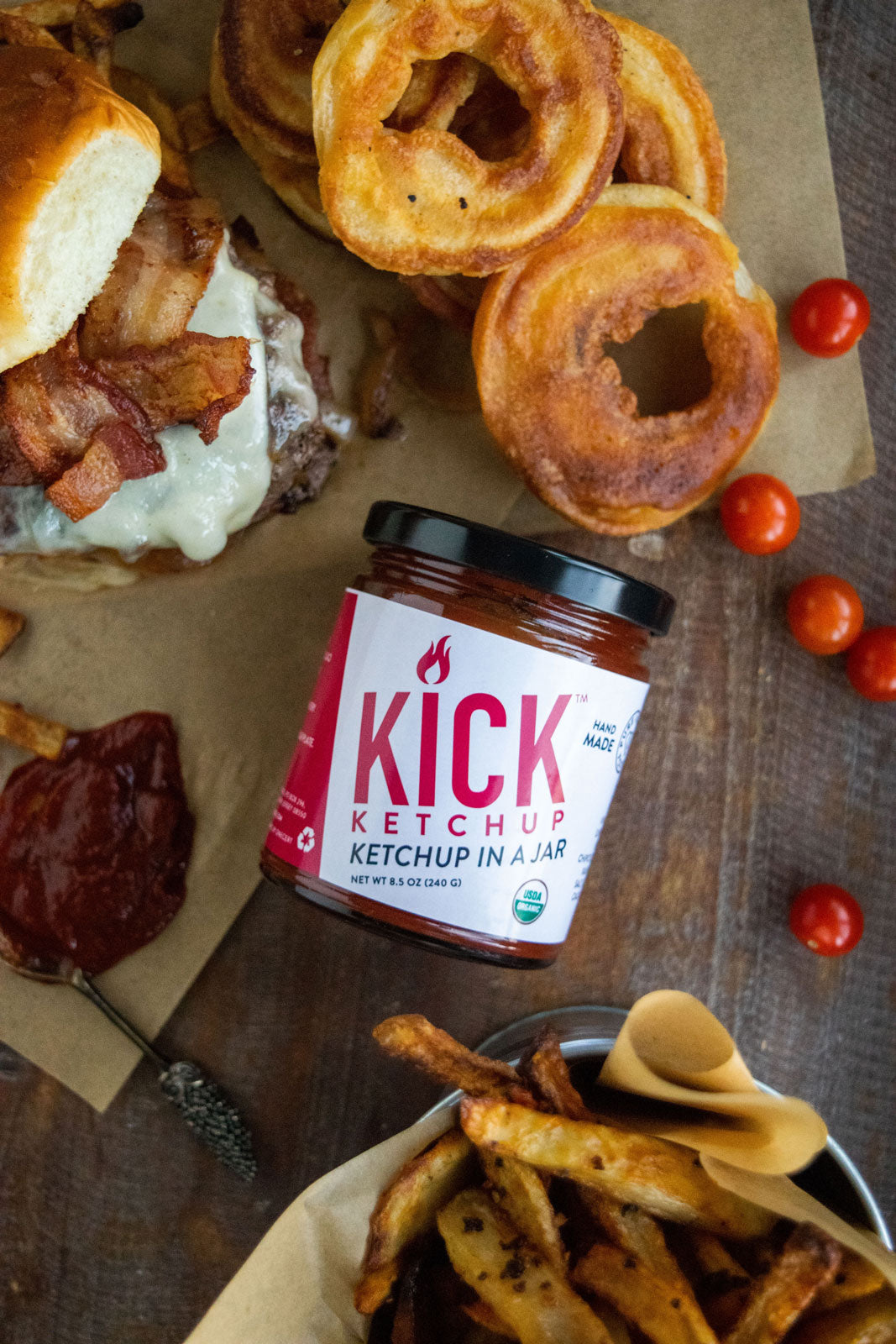 A jar of spicy ketchup from Pure Indian Foods, next to a drool-worthy burger, handmade onion rings, and fries. Certified organic.