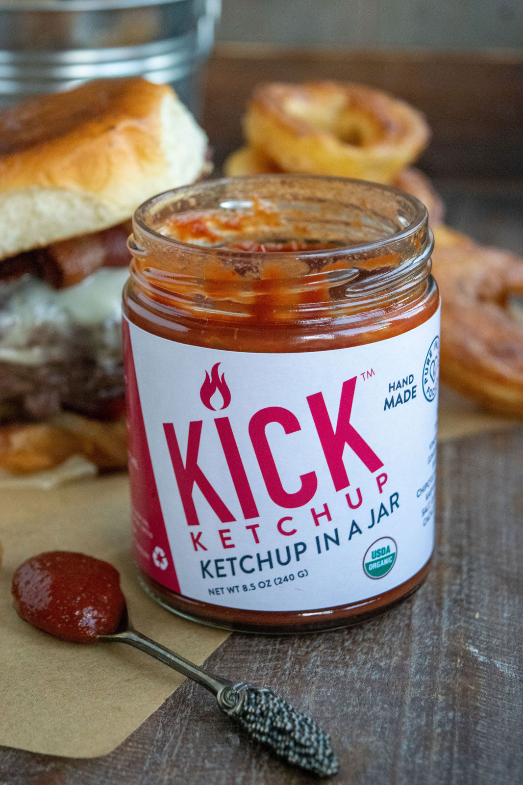 1 open jar of Pure Indian Foods KICK Ketchup, an organic ketchup. A heaping spoonful of ketchup is to the left of the jar, and a yummy looking burger with homemade onion rings is behind it.