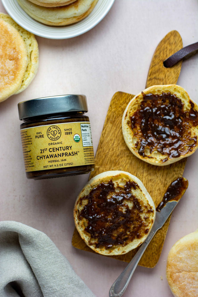 Jar of Pure Indian Foods organic chyawanprash jam, spread on a halved English muffin, displayed on a board with a small butter knife.