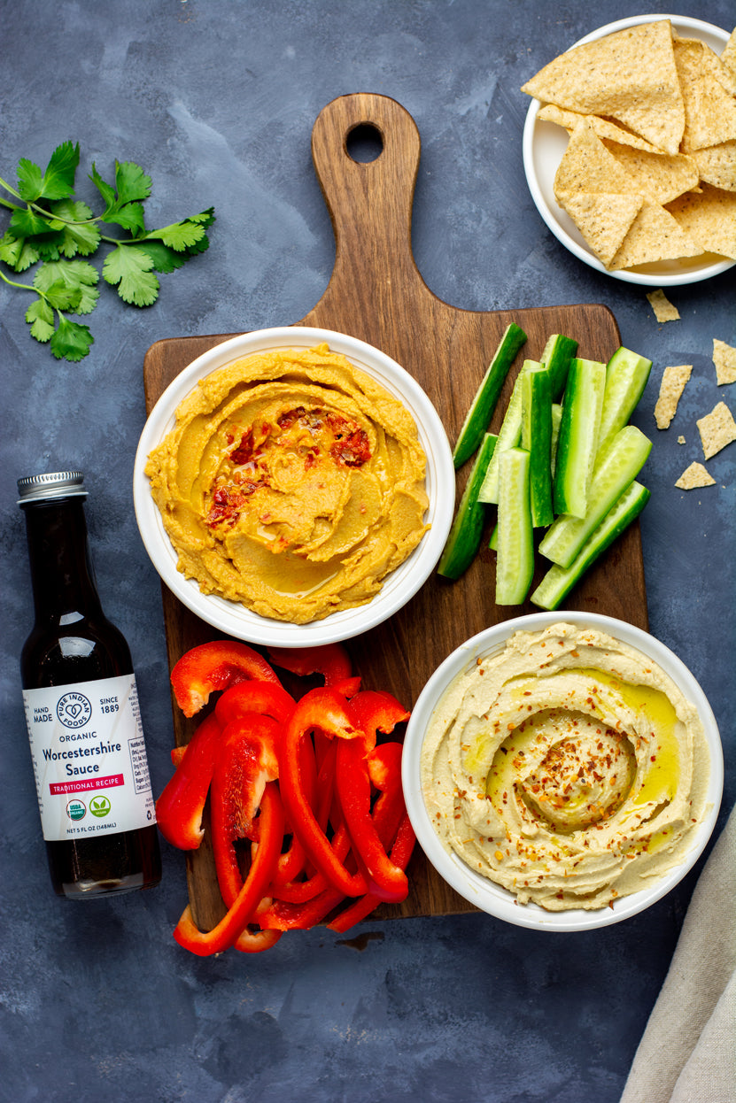 Hummus and dips on a serving board with freshly sliced veggies and chips, staged next to a jar of vegan Worcestershire sauce from Pure Indian Foods.
