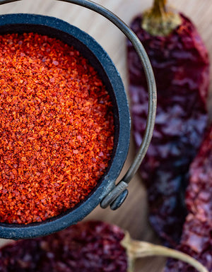 A small, black cast iron bowl filled with vibrant red ground cayenne pepper with dried peppers in the background