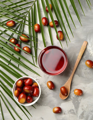 A clear container of red palm oil artfully displayed with palm leaves, a wooden spoon, and palm fruit